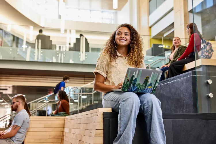 Student recognizes another student walk into the Belknap Academic Building as she is typing on her laptop and sitting on the wooden platform benches at the entrance of the BAB.