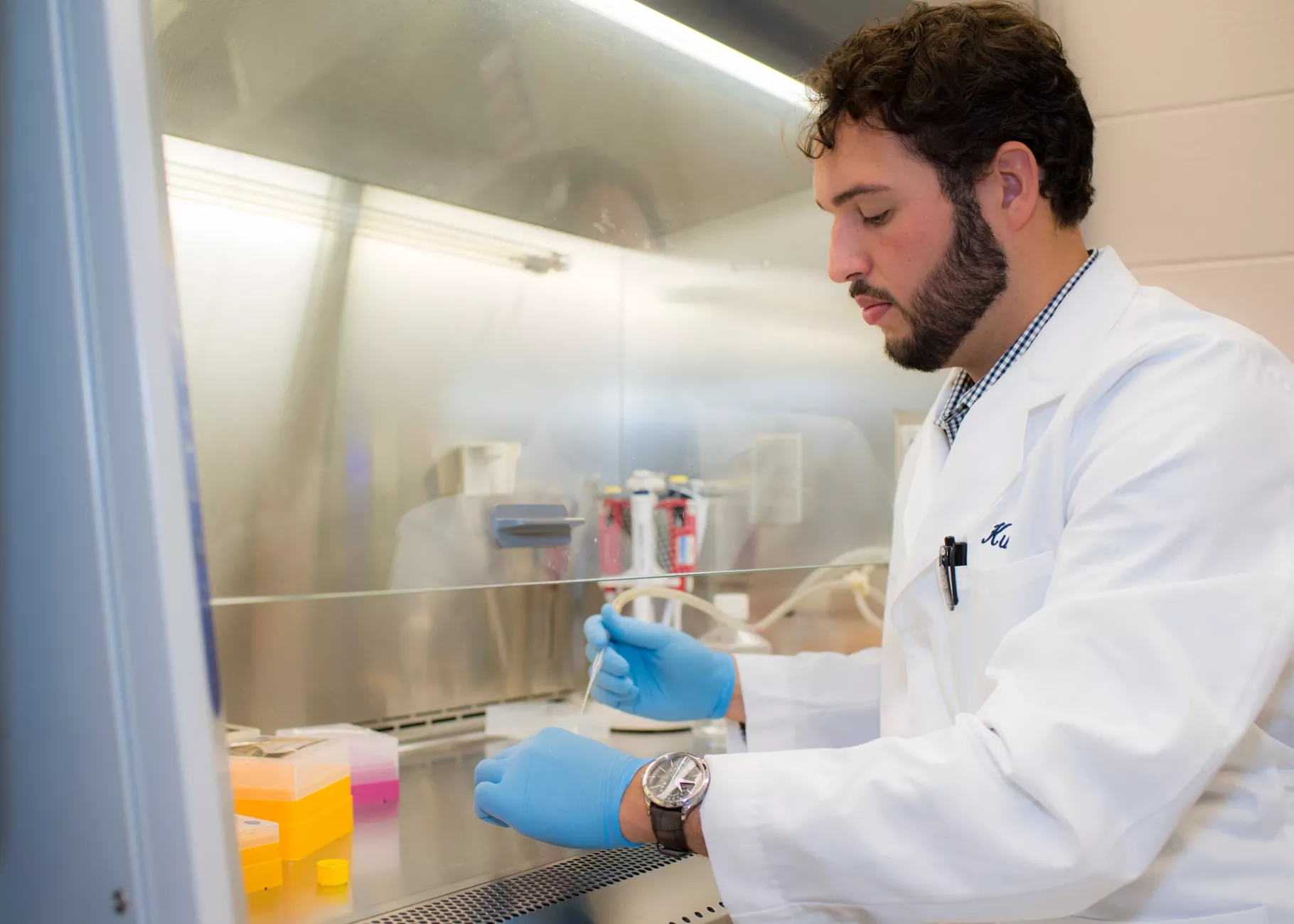 A man in a lab coat and gloves working with a pipette in a laboratory fume hood.