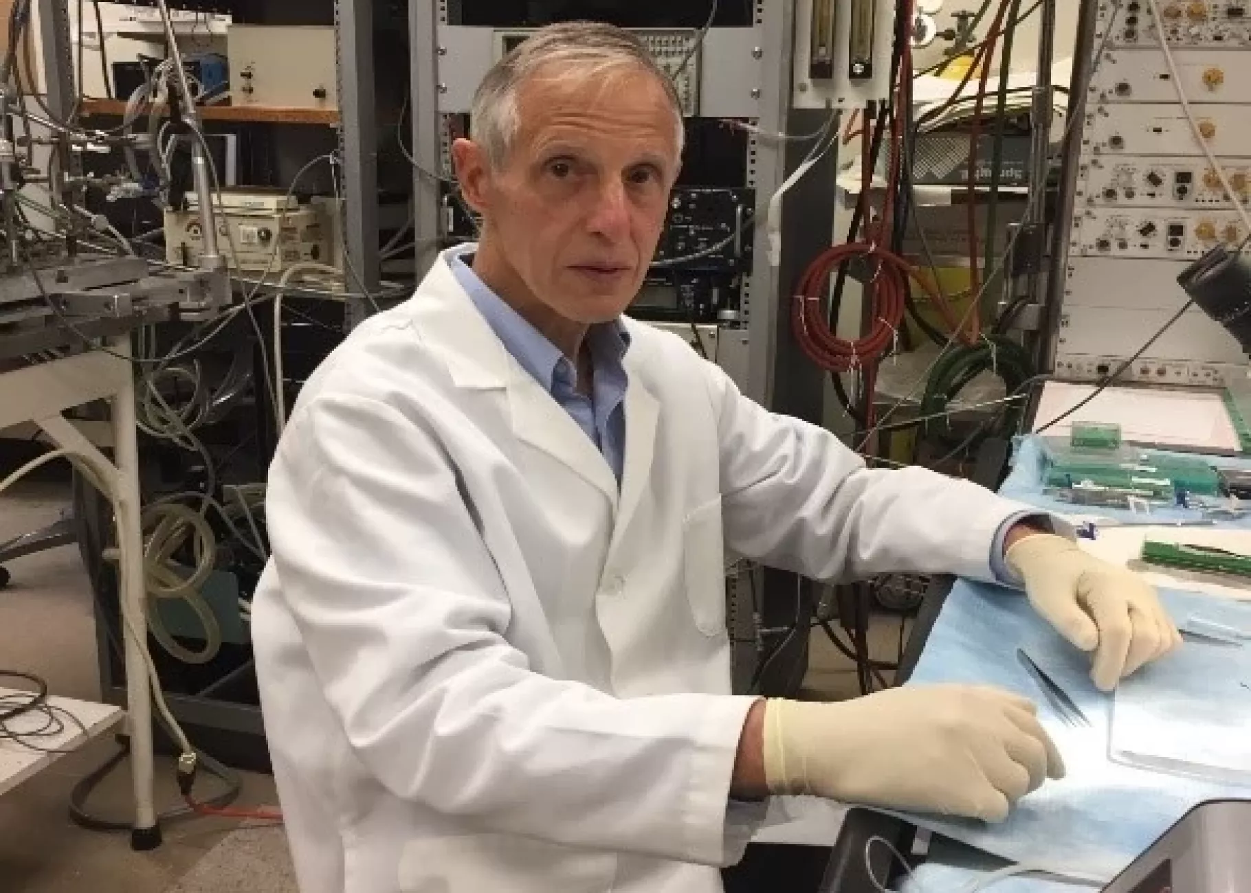 A person in a lab coat seated in a laboratory surrounded by electronic equipment.
