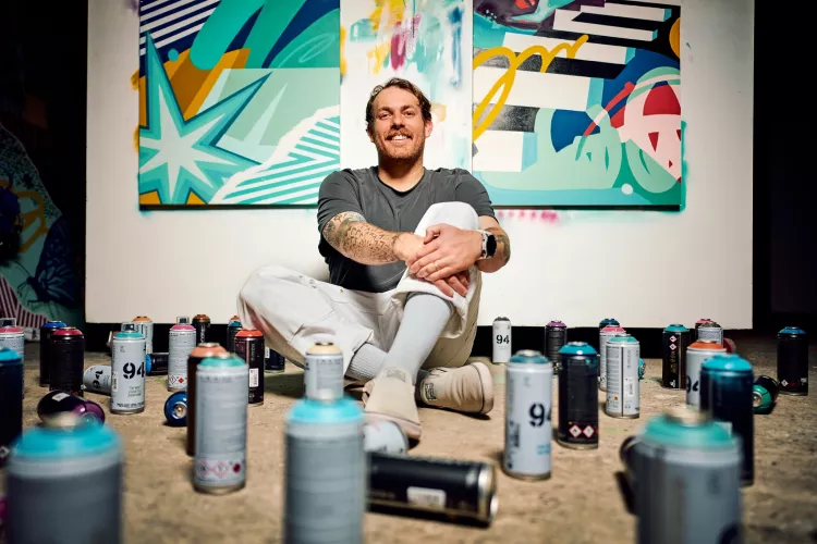 A smiling young man seated against a painted wall in his studio, surrounded by spray paint cans