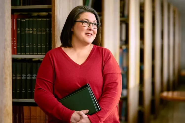 A smiling woman with glasses and holding books stands among shelves of library books.