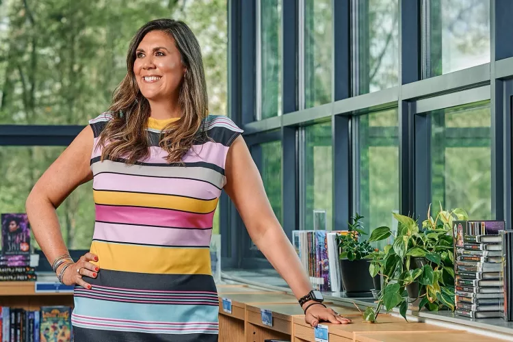 A smiling woman in a striped dress stands in an elementary school library.