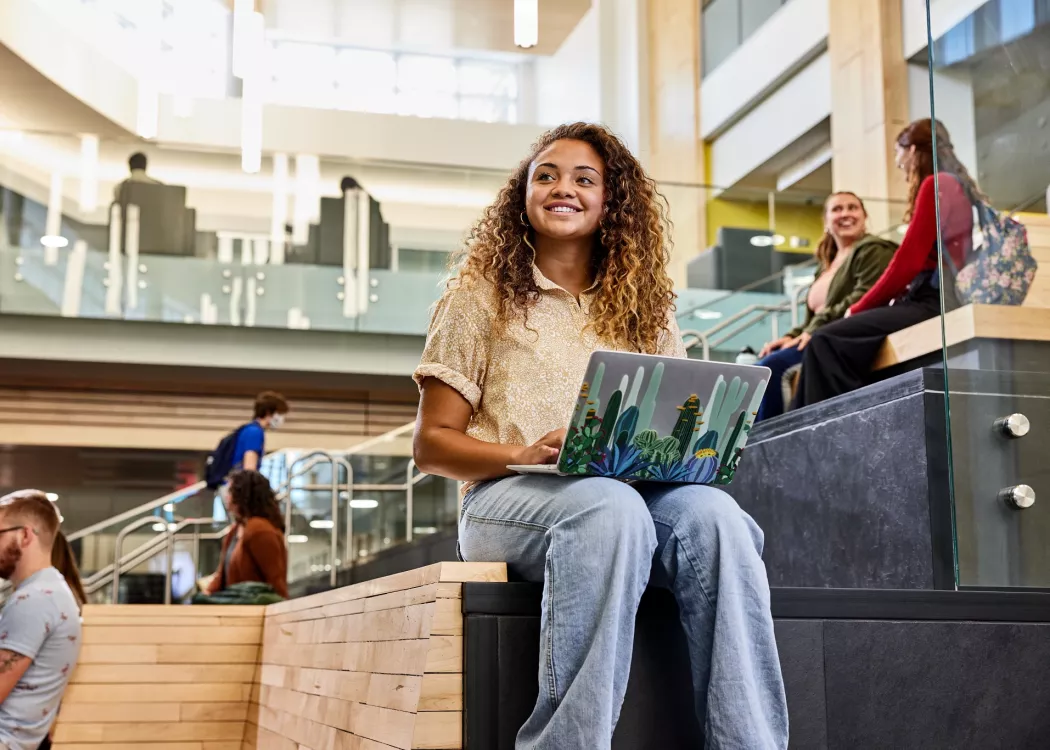 Student recognizes another student walk into the Belknap Academic Building as she is typing on her laptop and sitting on the wooden platform benches at the entrance of the BAB.