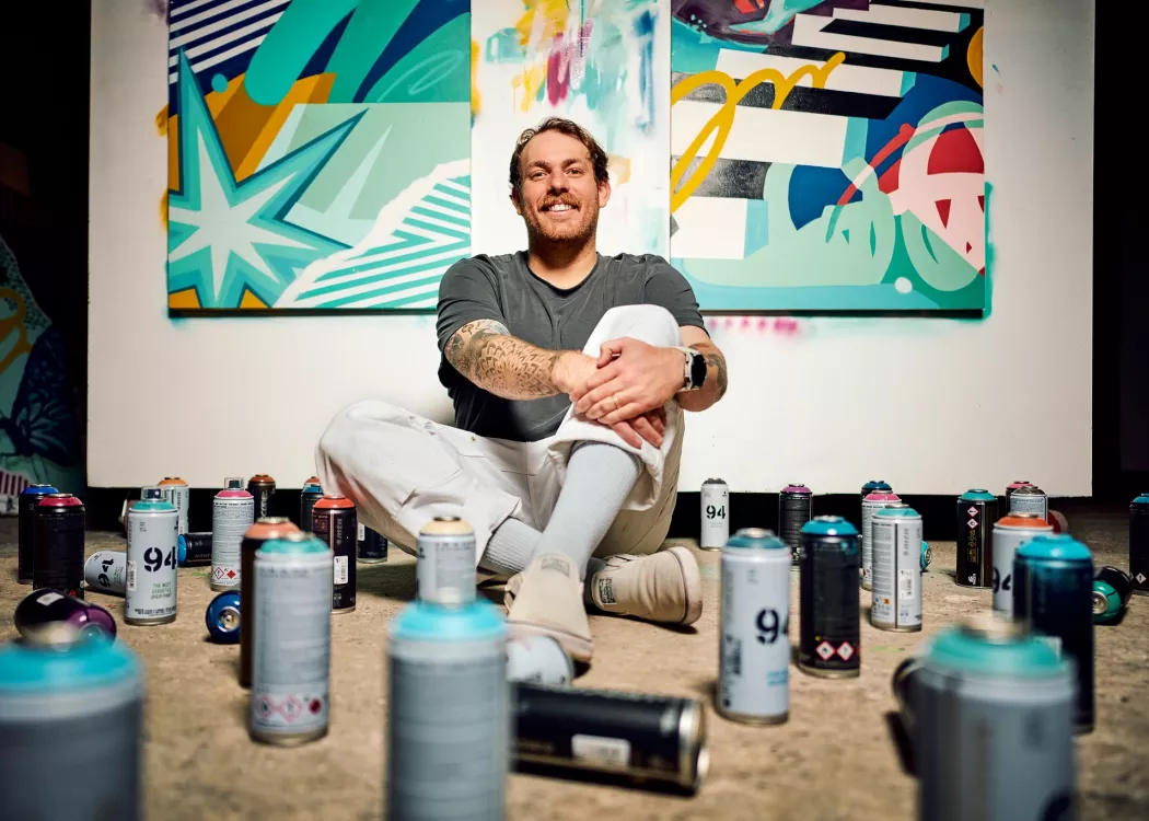 A smiling young man seated against a painted wall in his studio, surrounded by spray paint cans