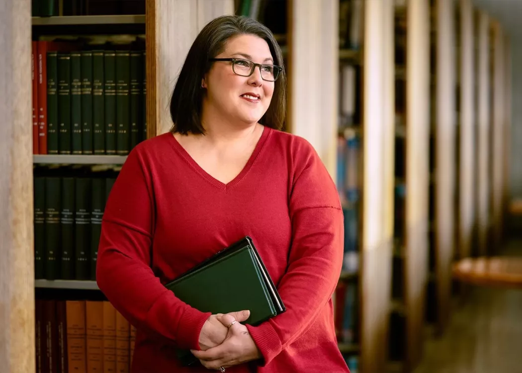 A smiling woman with glasses and holding books stands among shelves of library books.