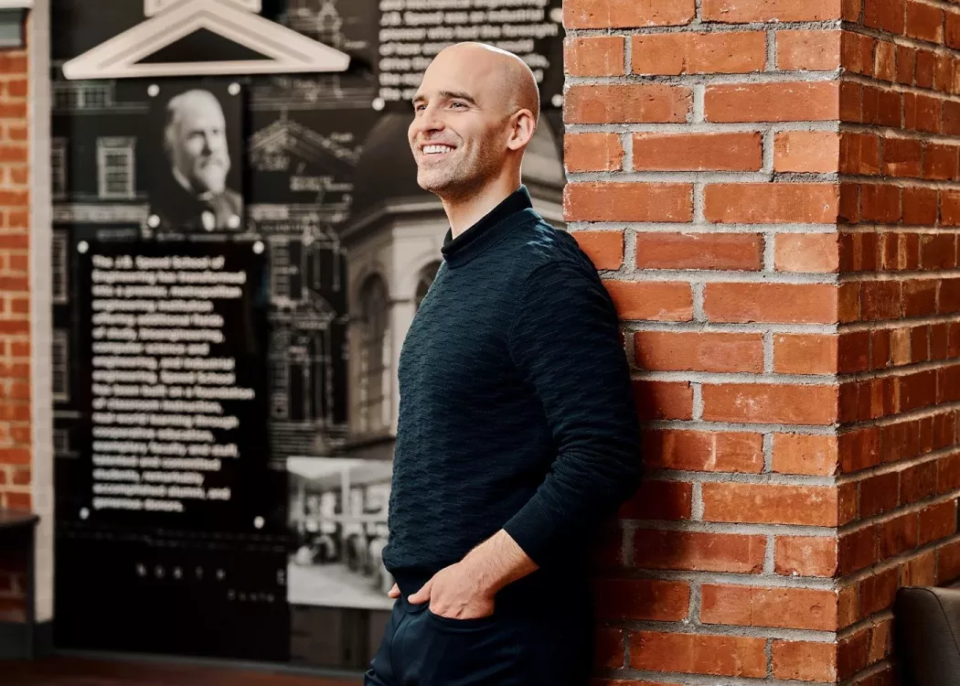 A smiling man in a black sweater poses against an industrial brick wall.