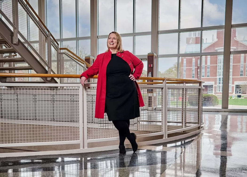 A smiling woman in a red jacket and black dress poses against a stairway railing.