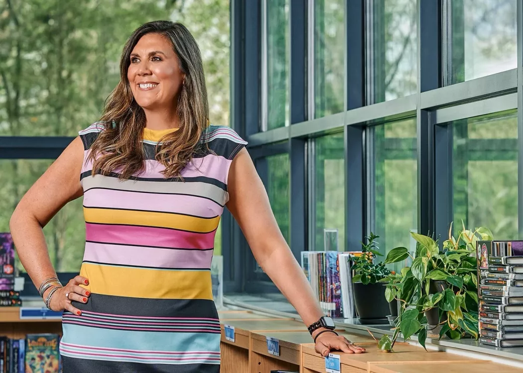 A smiling woman in a striped dress stands in an elementary school library.