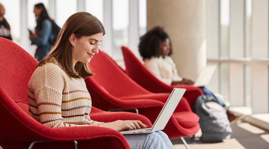 Lexi Raikes sitting on the second floor of the Belknap Academic Building, in a red chair, typing happily on her laptop