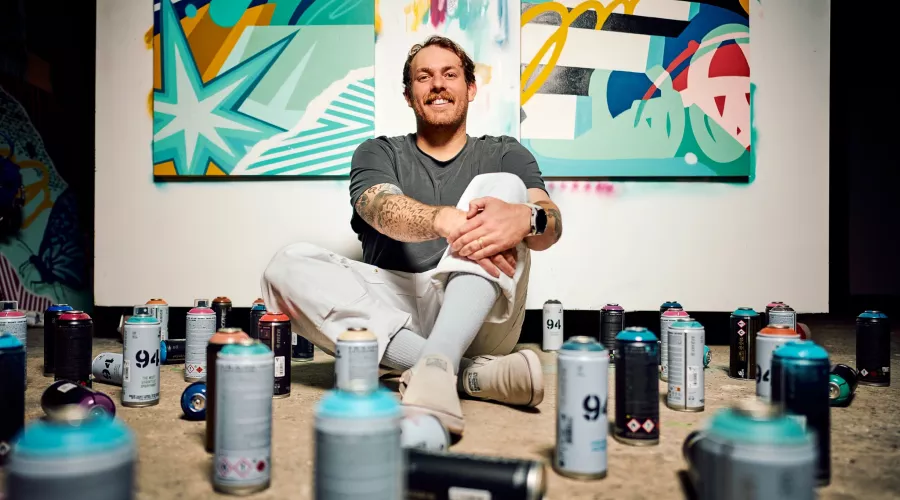 A smiling young man seated against a painted wall in his studio, surrounded by spray paint cans