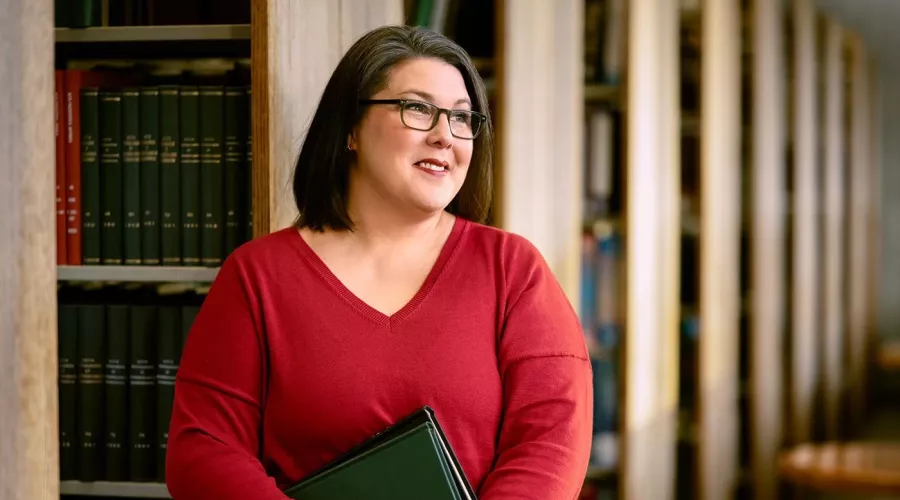 A smiling woman with glasses and holding books stands among shelves of library books.
