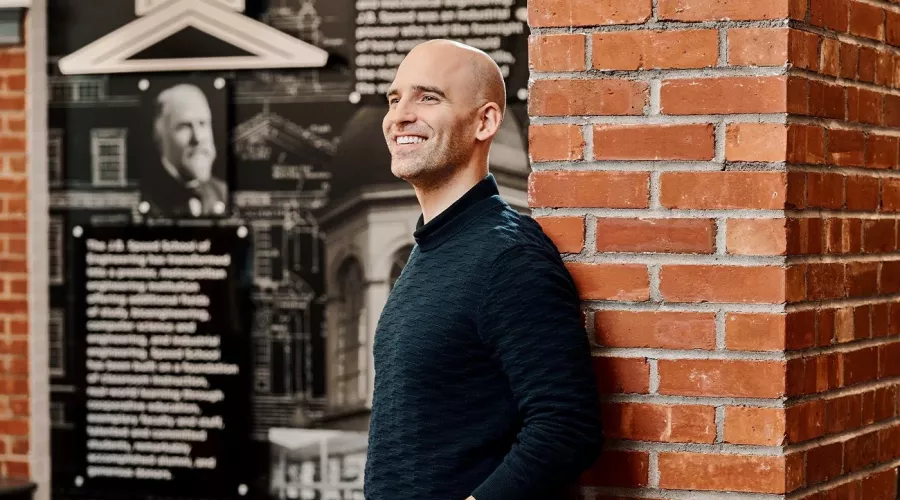 A smiling man in a black sweater poses against an industrial brick wall.