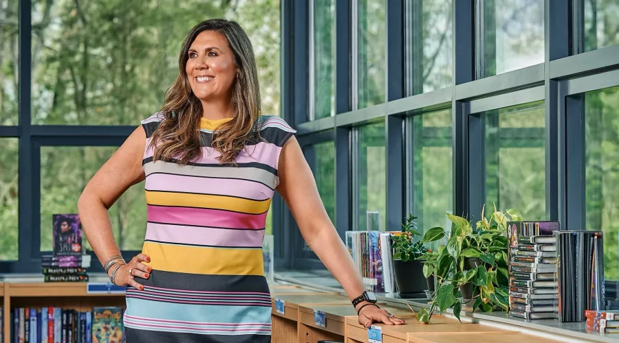 A smiling woman in a striped dress stands in an elementary school library.